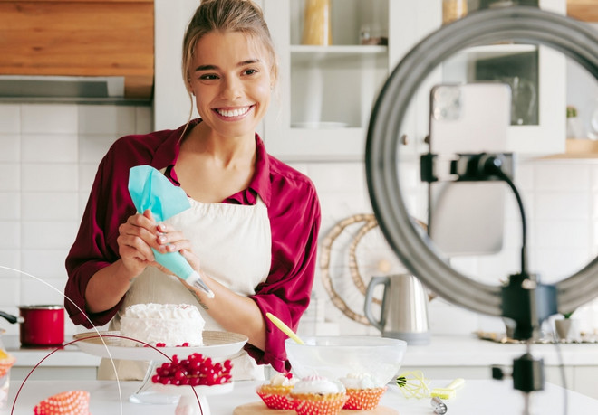 Eine blonde junge Frau verteilt Sahne auf einer Torte und lächelt dabei in ihre Handykamera, die vor einem Ringlicht befestigt ist
