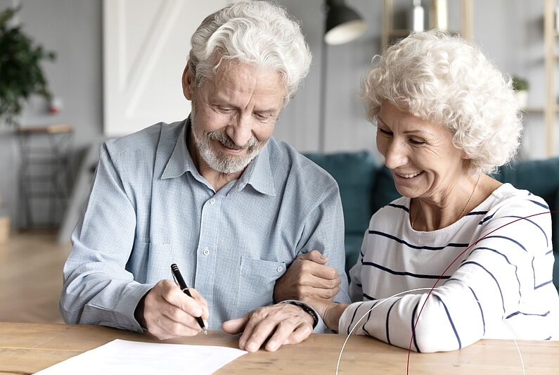 Ein älteres Ehepaar sitzt an einem Tisch. Der Mann unterschreibt gerade ein Dokument.