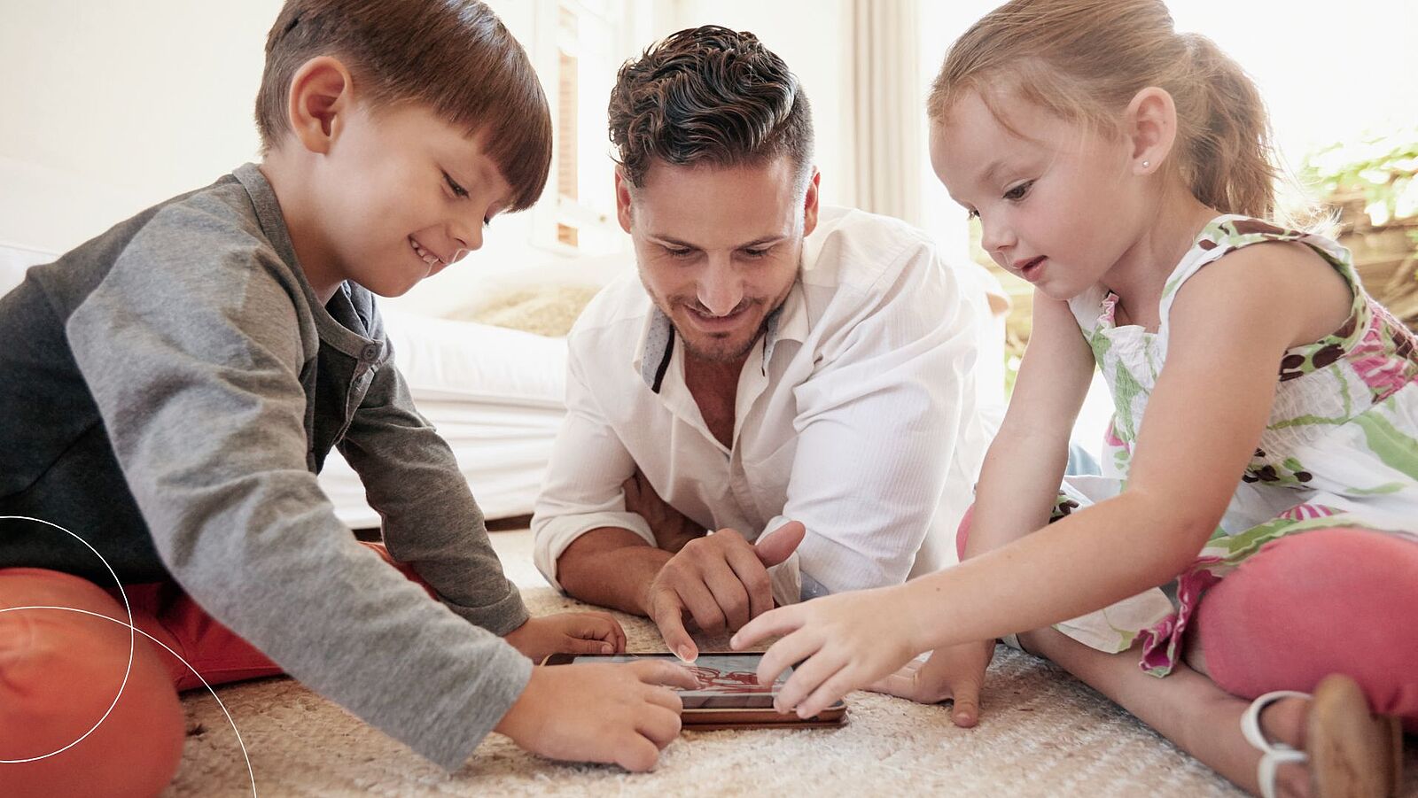 Ein Mann in der Mitte und ein Junge und ein Mädchen liegen auf dem Teppichboden und spiele mit einem Tablet.