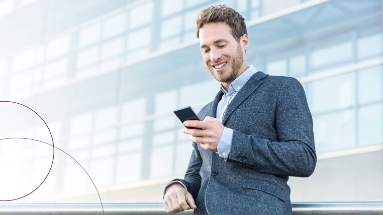 Ein junger Mann steht mit seinem Handy vor einem Bürogebäude und lächelt auf sein Handy herunter.